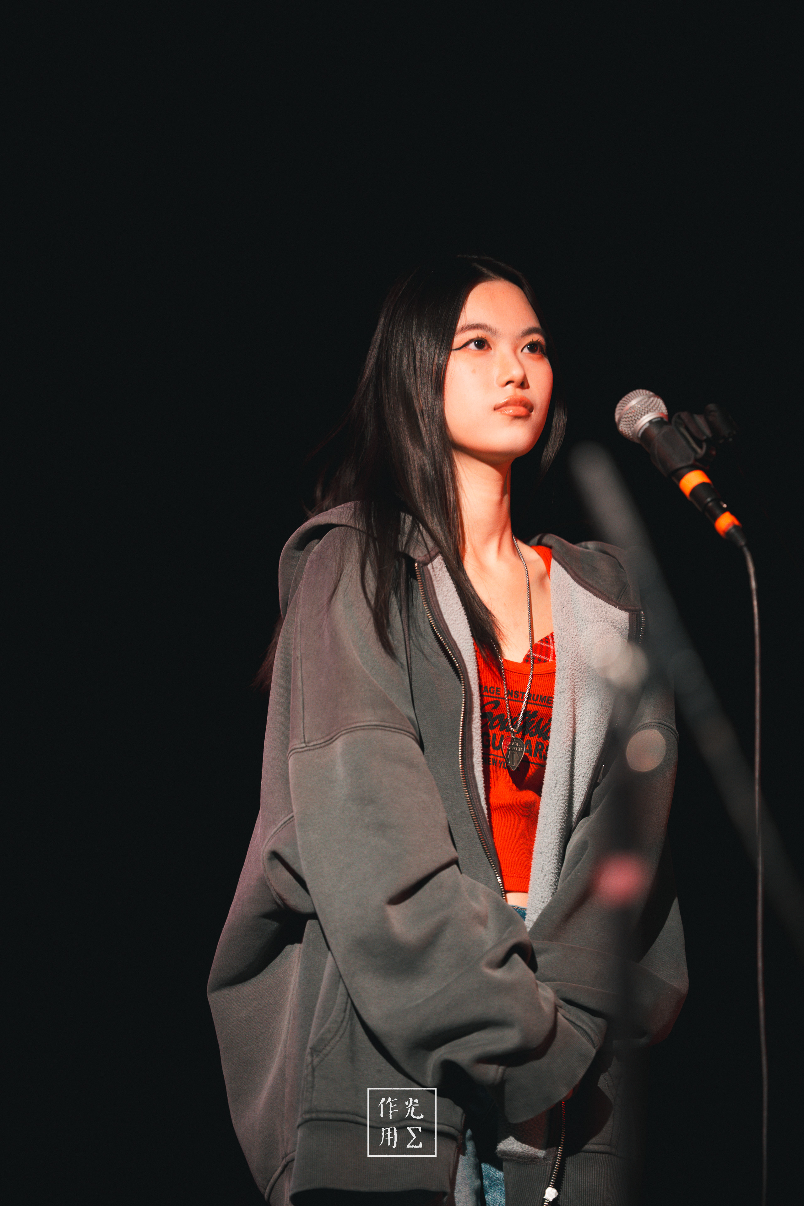 A young singer stands quietly beside a microphone on a dark stage