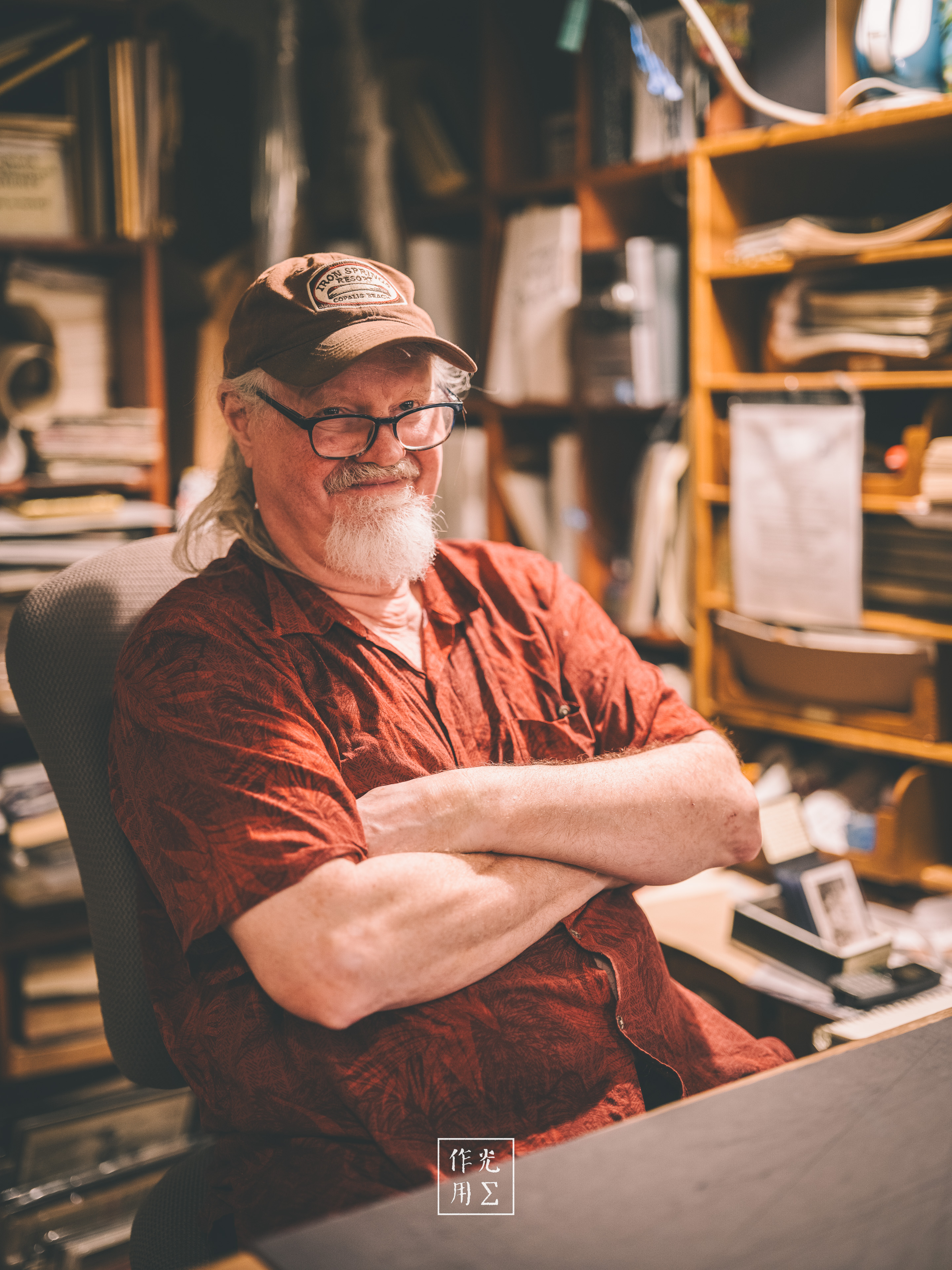 An older man in a cap sits with folded arms in a cluttered workshop