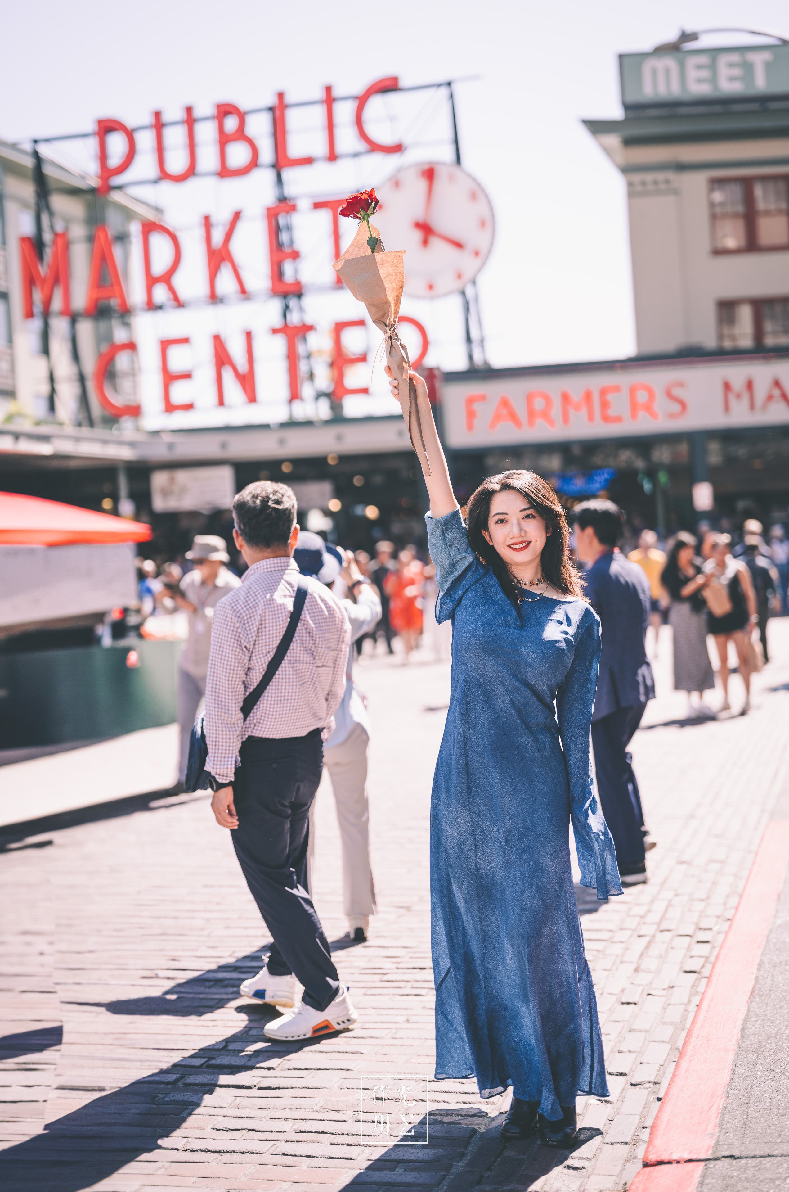 A woman in a long denim dress walks beneath the Public Market Center sign on a sunny busy street