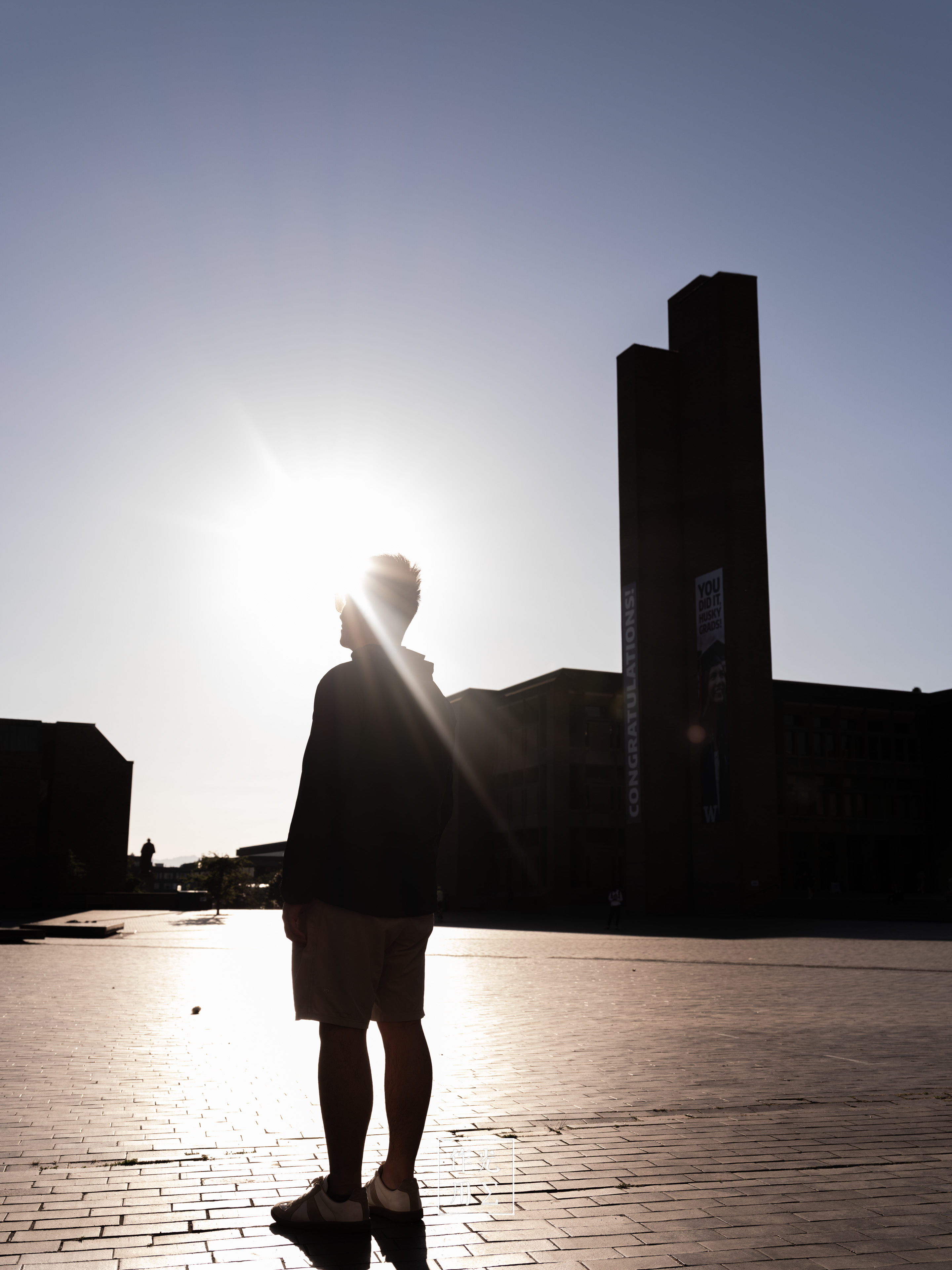 A silhouetted person stands on a sunlit plaza beside a tall modern tower