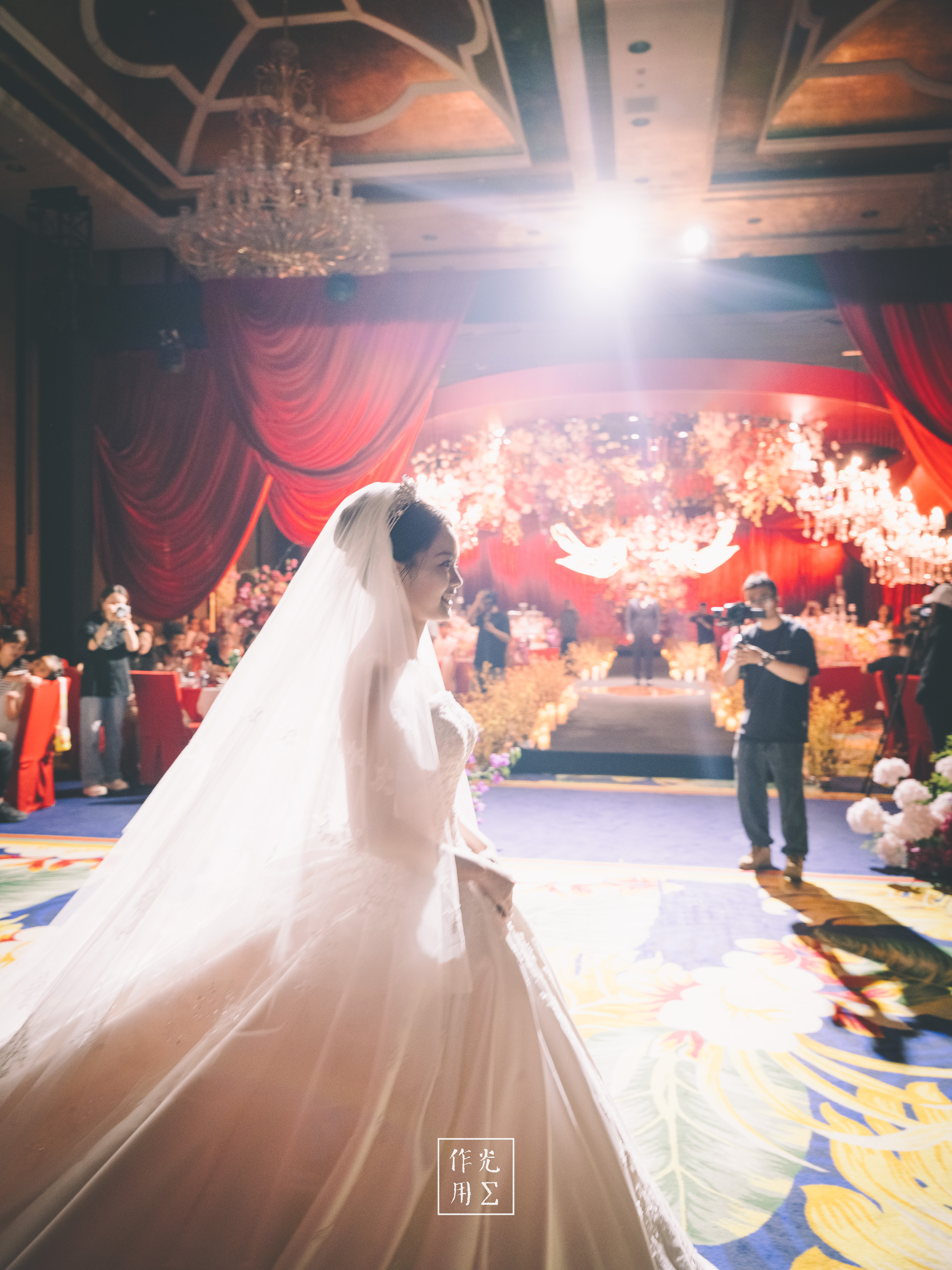 A veiled bride walks down a flower-lined wedding aisle beneath brilliant backlight